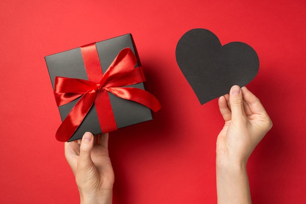 First person top view photo of hands holding black paper heart and black giftbox with red ribbon bow on isolated red background with copyspace