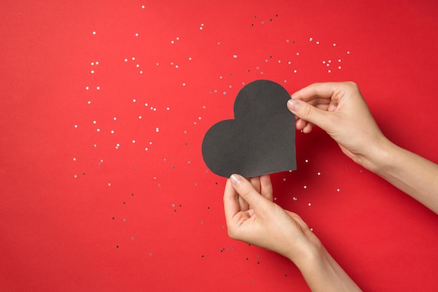 First person top view photo of hands holding black paper heart over shiny sequins on isolated red background with copyspace