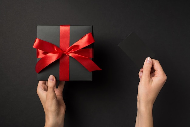 First person top view photo of hands holding black plastic card and black giftbox with red ribbon bow on isolated black background with empty space