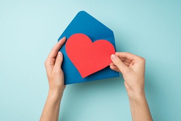 First person top view photo of hands holding open blue envelope with red paper heart on isolated pastel blue background with blank space