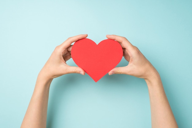 First person top view photo of hands holding red paper heart on isolated pastel blue background with blank space