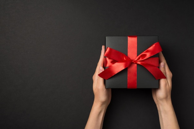 First person top view photo of hands holding stylish black giftbox with red satin ribbon bow on isolated black background with copyspace