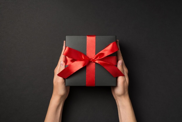 First person top view photo of hands holding stylish black giftbox with red satin ribbon bow on isolated black background