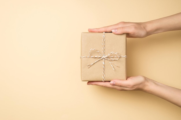 First person top view photo of hands holding stylish craft paper giftbox with twine bow on isolated beige background with blank space