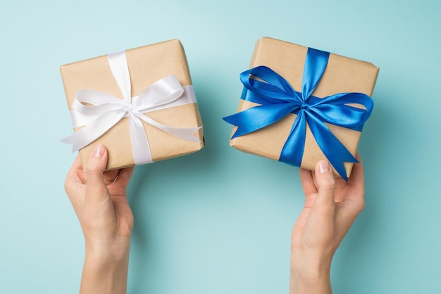 First person top view photo of hands holding two craft paper gift boxes with vivid blue and white ribbon bow on isolated pastel blue background