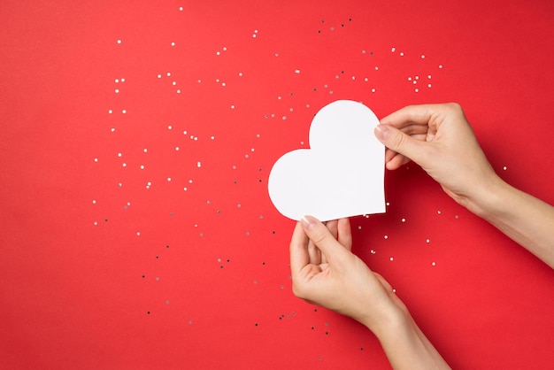 First person top view photo of hands holding white paper heart over shiny sequins on isolated red background with empty space