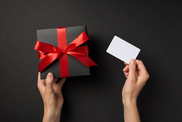 First person top view photo of hands holding white plastic card and black giftbox with red ribbon bow on isolated black background with blank space