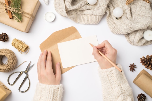 First person top view photo of hands in sweater writing letter giftbox scarf christmas tree balls handicraft tools cones anise candles scissors spool of twine isolated white background with copyspace