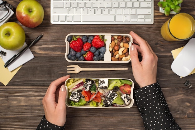 First person top view photo of hands touching two lunchboxes with salad berries and nuts fork apples glass of juice plant stationery keyboard and mouse on isolated dark wooden table background