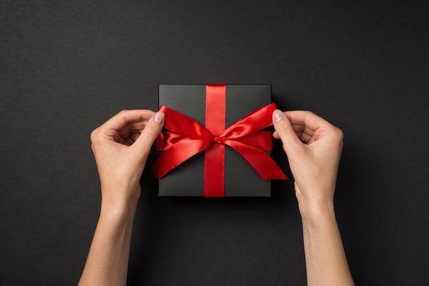 First person top view photo of hands tying red ribbon bow on black giftbox on isolated black background