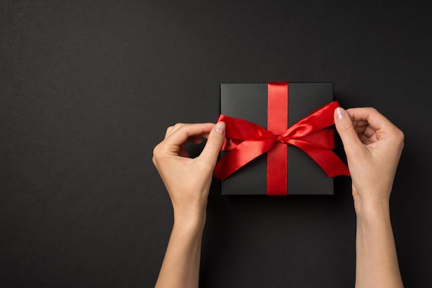 First person top view photo of hands tying vivid red satin ribbon bow on black giftbox on isolated black background with empty space