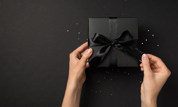 First person top view photo of hands unpacking black giftbox with black ribbon bow and tag over sequins on isolated black background with empty space