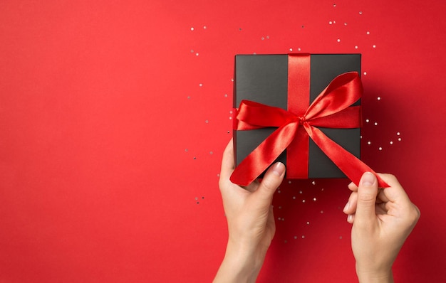 First person top view photo of hands unpacking black giftbox with red ribbon bow on isolated red background with blank space