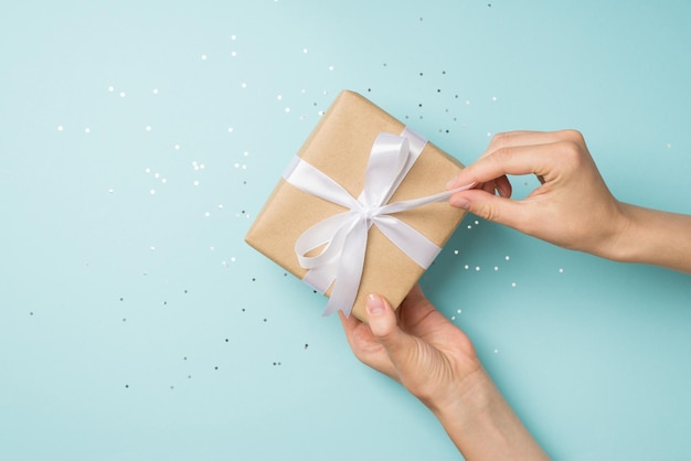 First person top view photo of hands unpacking craft paper gift box with white satin ribbon bow over sequins on isolated pastel blue background with copyspace