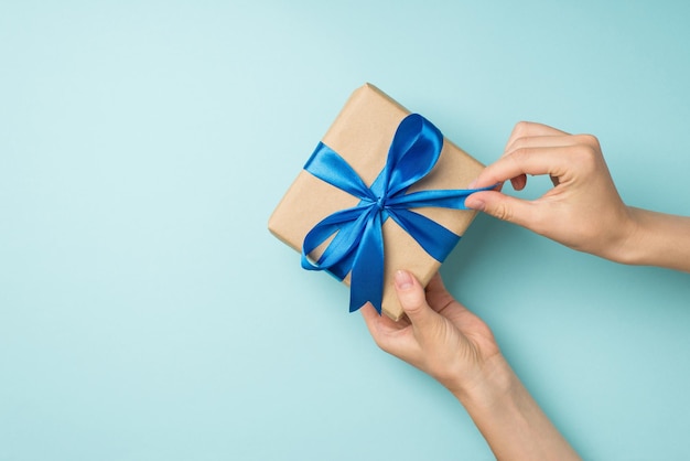 First person top view photo of hands unpacking craft paper giftbox with vivid blue satin ribbon bow on isolated pastel blue background with copyspace