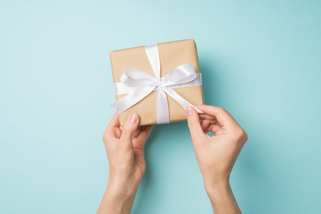 First person top view photo of hands unpacking craft paper giftbox with white ribbon bow on isolated pastel blue background