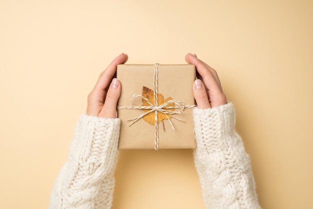 First person top view photo of hands in white sweater holding craft paper giftbox with twine bow and yellow autumn leaf on isolated beige background