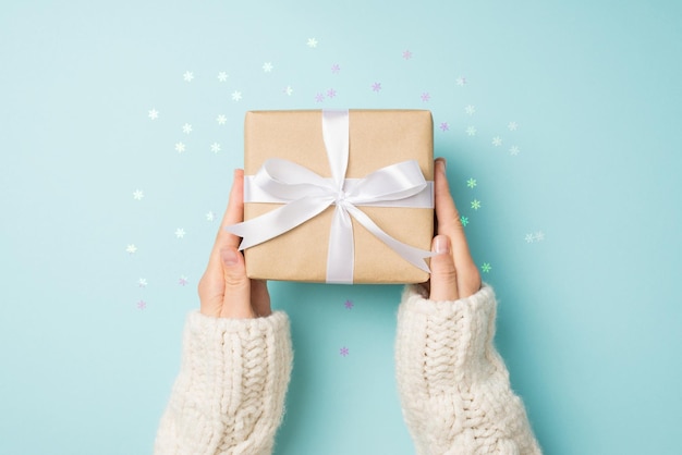 First person top view photo of hands in white sweater holding craft paper giftbox with white ribbon bow over shiny snowflakes confetti on isolated pastel blue background