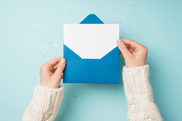 First person top view photo of hands in white sweater holding open blue envelope with white paper sheet over sequins on isolated pastel blue background with empty space