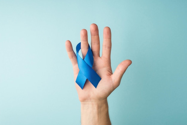 First person top view photo of male hand with blue ribbon on ring finger symbol of prostate cancer awareness on isolated pastel blue background