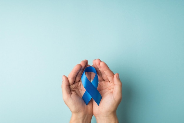 First person top view photo of man's hands holding blue ribbon in palms symbol of prostate cancer awareness on isolated pastel blue background with copyspace