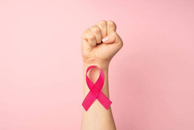 First person top view photo of raised female hand with clenched fist and pink ribbon on wrist symbol of breast cancer awareness on isolated pastel pink background with copyspace