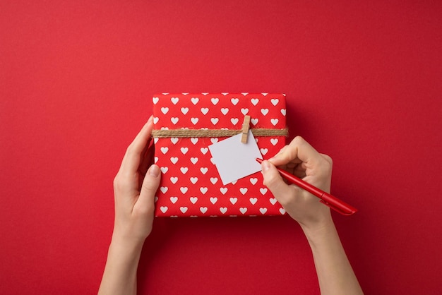 First person top view photo of saint valentine's day decor girl's hands writing note attached to giftbox in red wrapping paper with heart pattern and twine on isolated red background with copyspace