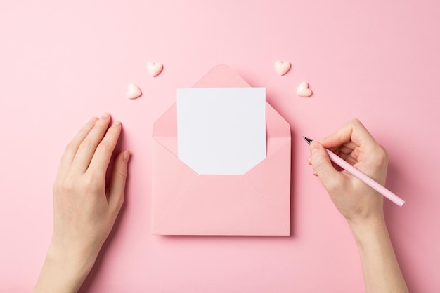 First person top view photo of saint valentine's day decorations girl's hands holding pen over open pink envelope with letter and pink hearts on isolated pastel pink background with blank space