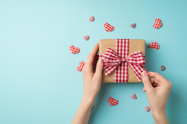 First person top view photo of st valentine's day decor hands untying checkered ribbon bow on kraft paper giftbox surrounded by decorative hearts on isolated pastel blue background with empty space