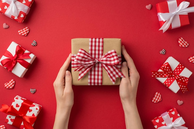 First person top view photo of valentine's day decor female hands holding craft paper giftbox with checkered ribbon bow hearts white and red present boxes on isolated red background