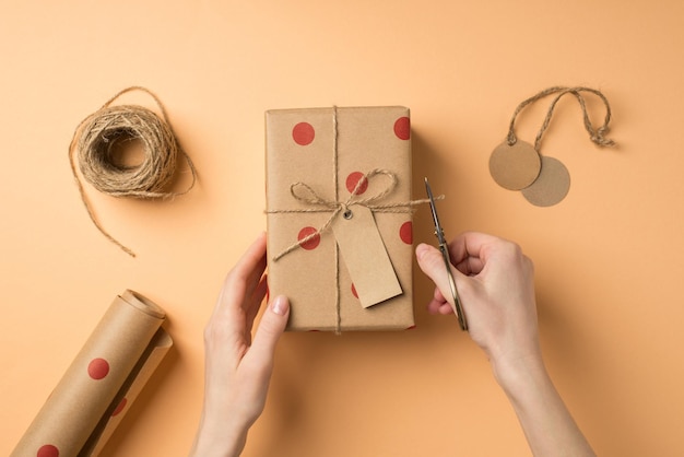 First person top view photo of valentine's day decor handicraft tools girl's hands cutting twine on craft paper giftbox with polka dot pattern label bow on isolated beige background with empty space