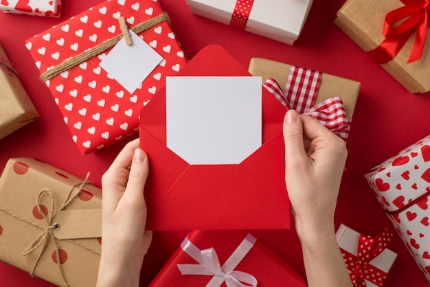 First person top view photo of valentine's day decor young girl's hands holding open red envelope with card over present boxes on isolated red background with blank space