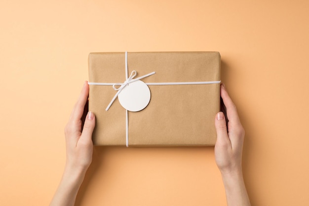 First person top view photo of valentine's day decorations young woman's hands holding kraft paper giftbox with small bow and pricetag on isolated beige background with copyspace