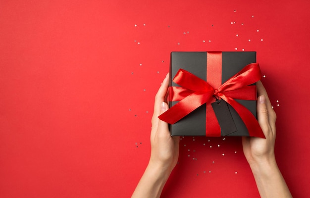 First person top view photo of woman hands holding black giftbox with red ribbon bow and tag sequins on isolated red background with copyspace