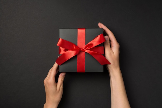 First person top view photo of woman hands touching stylish black giftbox with red satin ribbon bow on isolated black background