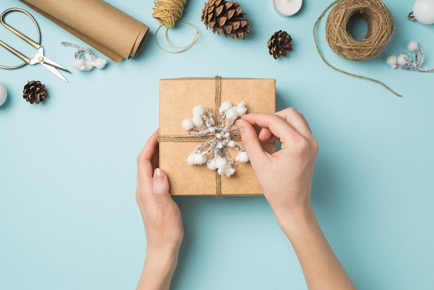 First person top view photo of woman's hands decorating gift box with snow twig and twine over white christmas tree balls and handicraft tools on isolated pastel blue background