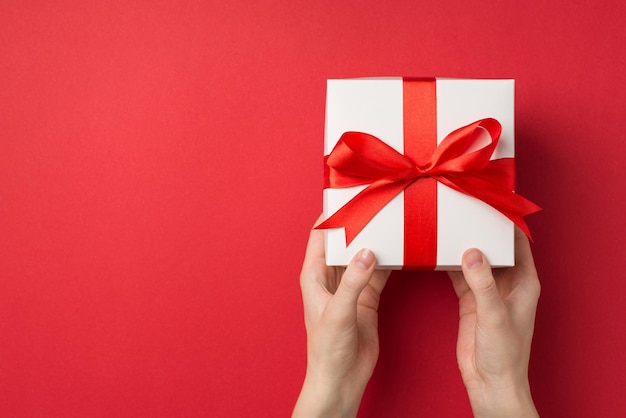 First person top view photo of woman's hands giving big white giftbox with red silk ribbon bow on isolated red background with blank space