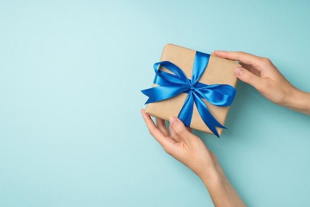 First person top view photo of woman's hands holding craft paper giftbox with blue satin ribbon bow on isolated pastel blue background with copyspace