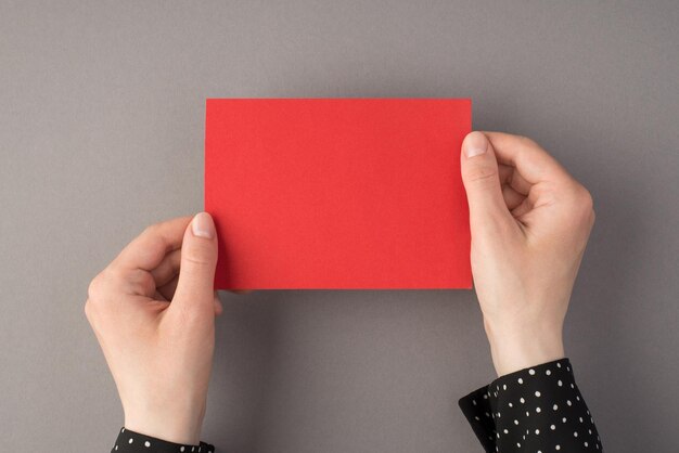 First person top view photo of woman's hands holding red paper card on isolated grey background with copyspace