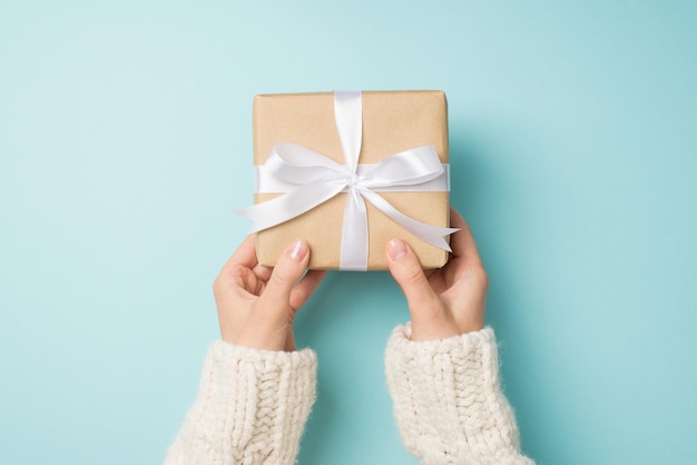 First person top view photo of woman's hands in white cozy sweater holding craft paper giftbox with white satin ribbon bow on isolated pastel blue background