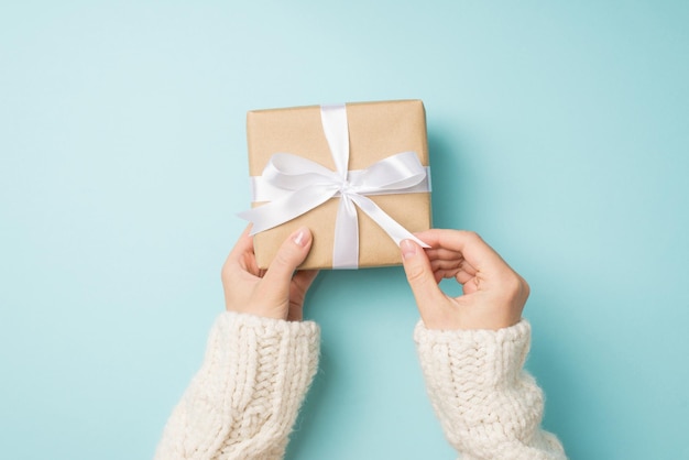 First person top view photo of woman's hands in white cozy sweater unpacking craft paper giftbox with white satin ribbon bow on isolated pastel blue background