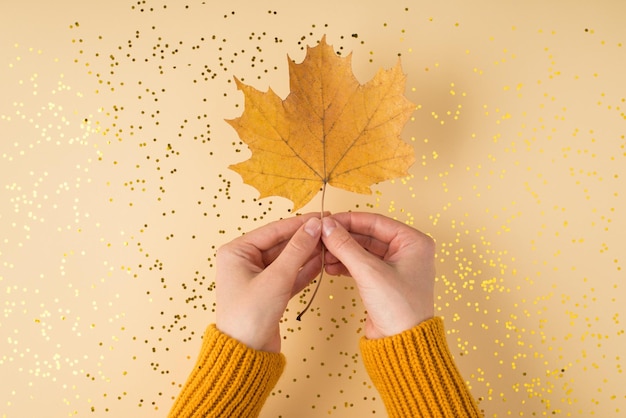 First person top view photo of woman's hands in yellow pullover holding orange autumn maple leaf over scattered golden sequins on isolated pastel orange background