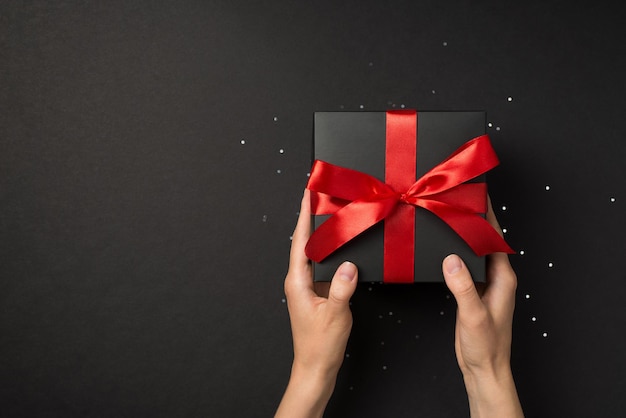 First person top view photo of young person hands holding black giftbox with vivid red ribbon bow over shiny sequins on isolated black background with blank space