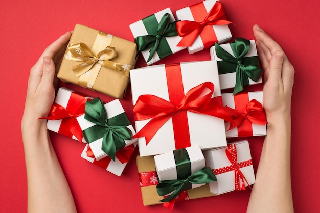 First person top view photo of young woman's hands hugging stack of gift boxes with satin ribbon bows on isolated red background