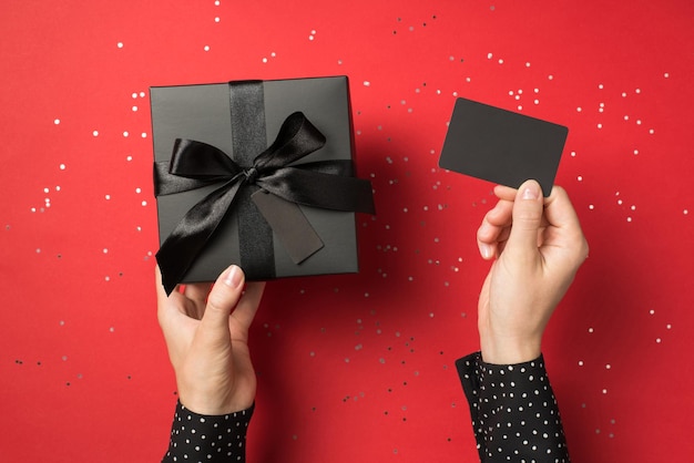 First person view overhead photo of black giftbox with ribbon wrapped as bow and business card in hands and confetti around isolated on the red background