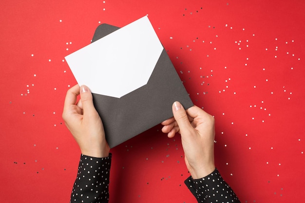 First person view overhead photo of woman hands holding black envelope with white card inside isolated on the red background with confetti