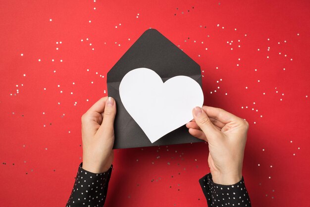 First person view above photo of female hands holding black envelope with white card as heart inside isolated on the red background with confetti