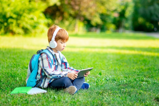 A firstgrader boy with a backpack and a tablet with headphones on a green lawn reading a book or doing homework going back to school