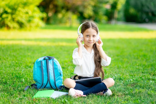 A firstgrader girl with a backpack and a tablet with headphones on a green lawn reads a book or does homework goes back to school