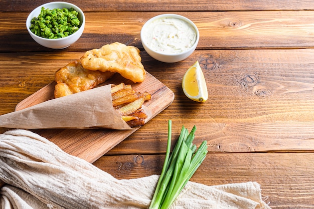 Fish & chips with dip and lemon, mashed minty peas, tartar sauce in paper cone  on wood chopping board dip and lemon - fried cod, french fries, lemon slices, tartar sauce.